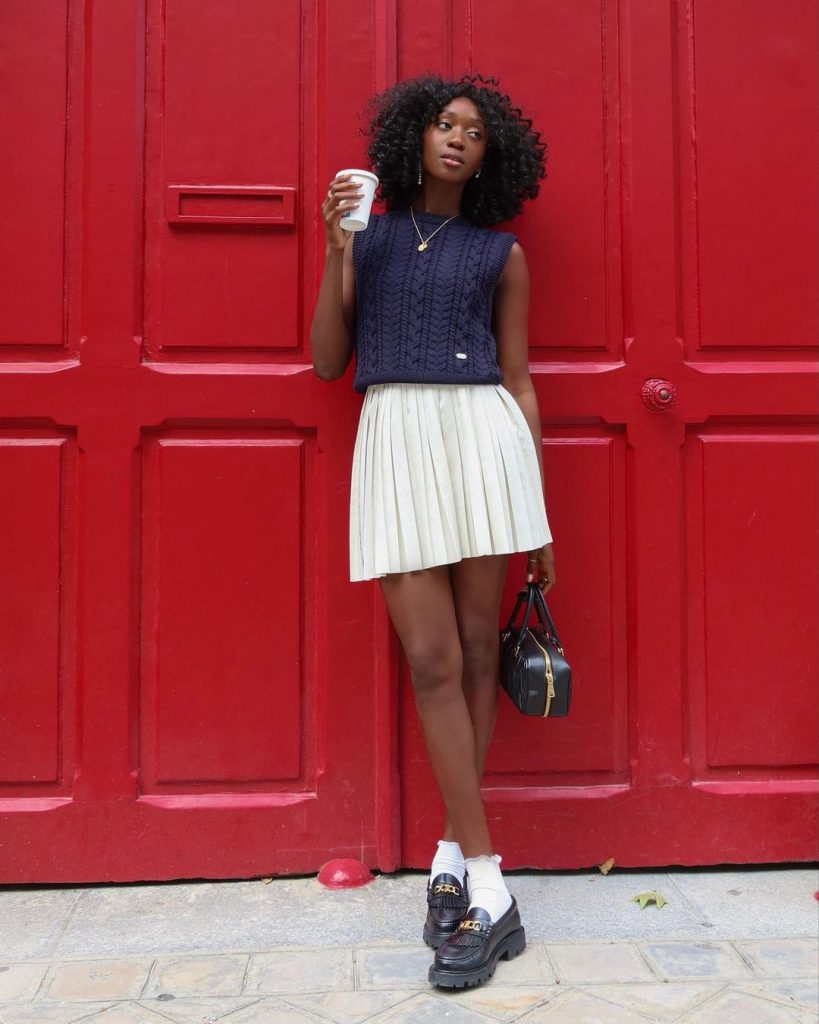 Emmanuelle Koffi com blusa de tricô, saia curta plissada, meia branca e mocassim