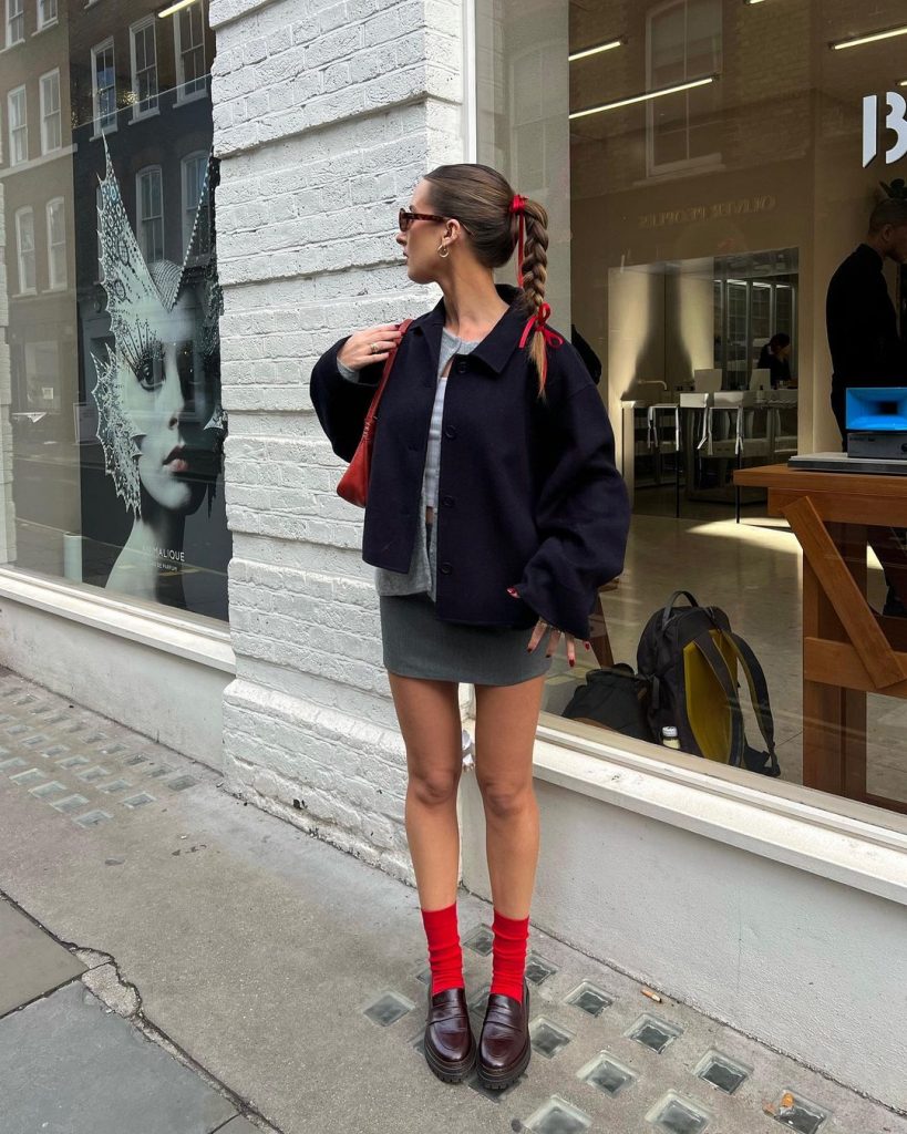 Kezia Rose Cook com la&ccedil;o de fita no cabelo, &oacute;culos de sol, casaco azul-marinho, cardigan cinza, regata cinza, minissaia cinza, meia vermelha e mocassim vinho