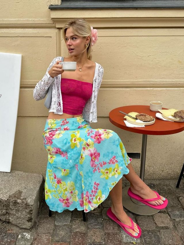 Linda com acessório de flor no cabelo, casaco de renda, blusa rosa sem alça, saia midi fluida estampada e Havaianas rosa